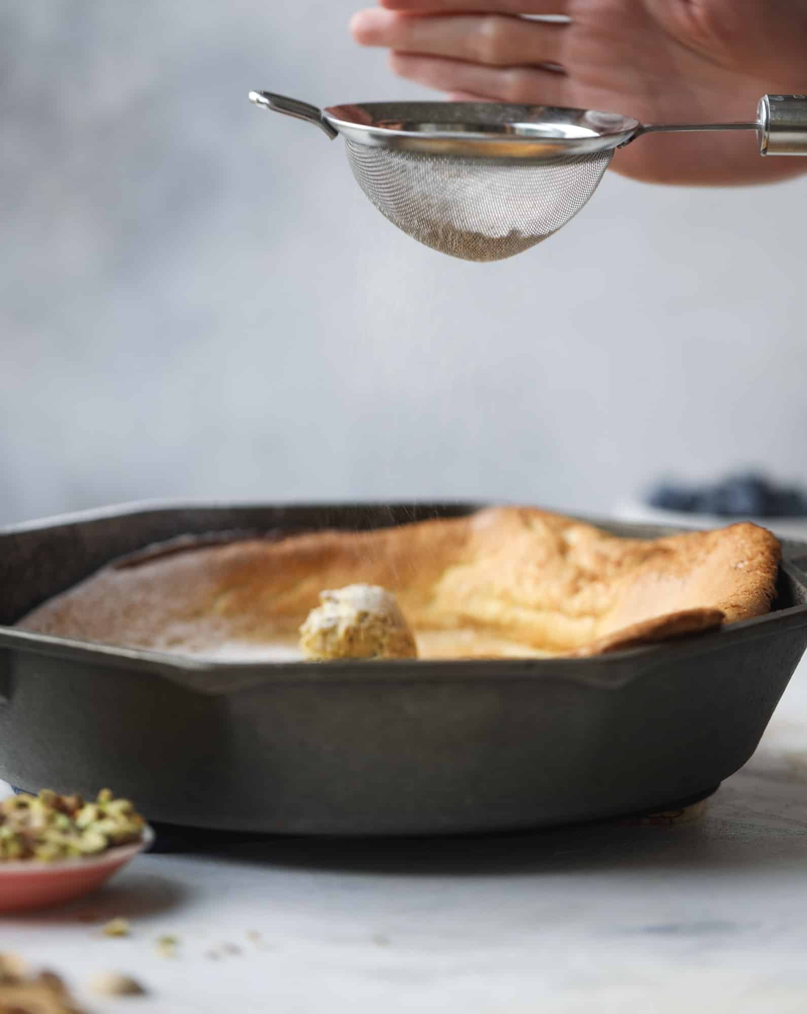 This incredible dutch baby recipe is fluffy and light and perfect for breakfast! It's sprinkled with pistachio sugar and served with pistachio butter and fresh blueberries. It's super easy, simple and always a hit! I howsweeteats.com #dutch #baby #pistachio #blueberry #breakfast
